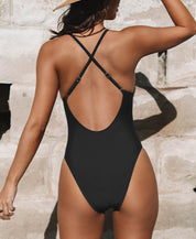 Donna in costume intero nero con spalline incrociate, sfondo di muro in pietra. Moda mare, abbigliamento estivo, tendenza costumi da bagno 2023.