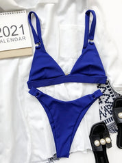 Bikini blu a due pezzi su sfondo bianco, con reggiseno triangolare e slip a vita alta. Accessori: sandali neri con perle, calendario 2021.