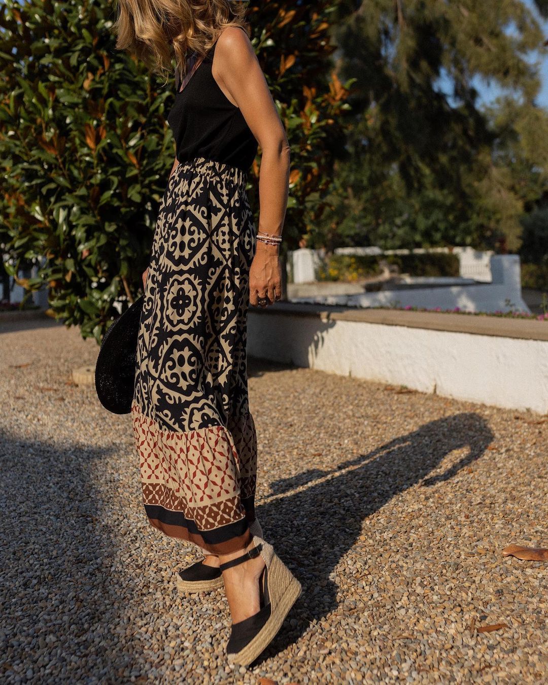 Donna con gonna lunga a motivi geometrici, top nero e espadrillas nere su ghiaia. Moda estiva, abbigliamento casual chic, stile boho.