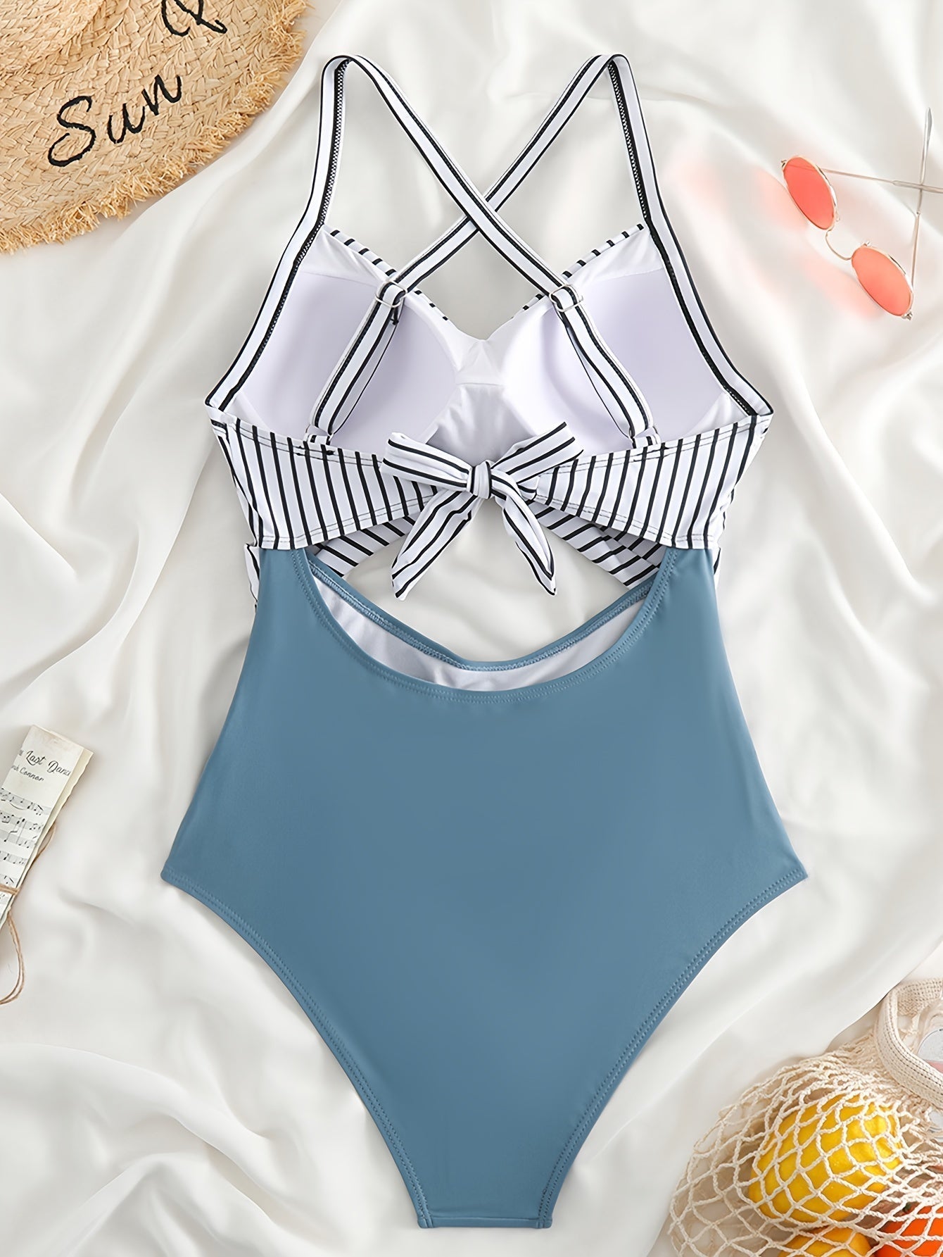 Costume intero donna a righe bianche e nere con fiocco, parte inferiore blu, su sfondo bianco con occhiali da sole e cappello di paglia.