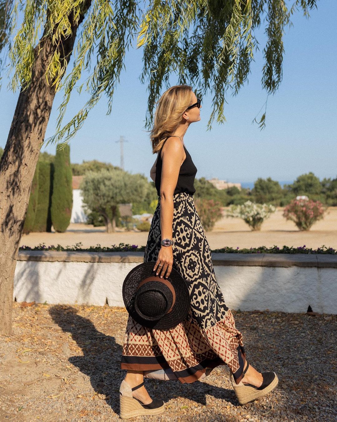 Donna con gonna lunga a motivi geometrici, top nero e sandali con zeppa, cammina all'aperto sotto un albero, tenendo un cappello di paglia.