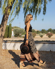 Donna con gonna lunga a motivi geometrici, top nero e sandali con zeppa, cammina all'aperto sotto un albero, tenendo un cappello di paglia.
