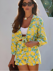 Donna con occhiali da sole indossa un completo estivo con stampa di limoni, giacca e pantaloncini, su sfondo bianco. Moda estiva, abbigliamento casual.