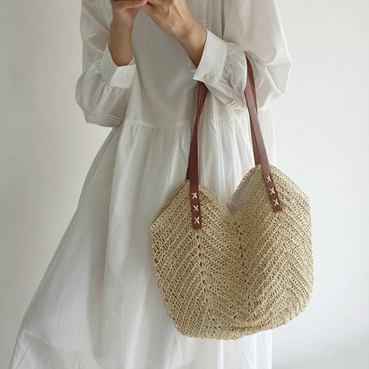 Donna con abito bianco tiene borsa di paglia intrecciata con manici in pelle marrone. Moda estiva, accessorio casual chic, tendenza 2023.