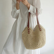 Donna con abito bianco tiene borsa di paglia intrecciata con manici in pelle marrone. Moda estiva, accessorio casual chic, tendenza 2023.