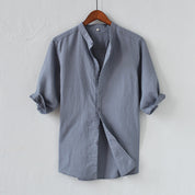 Camicia uomo grigia in cotone, maniche lunghe arrotolate, colletto alla coreana, design casual elegante, appesa su gruccia di legno.