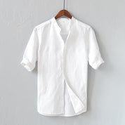 Camicia bianca in lino da uomo, maniche corte, colletto alla coreana, appesa su gruccia di legno. Elegante e casual, ideale per l'estate.