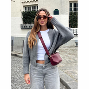 Donna con occhiali da sole, maglione grigio, top bianco e jeans, indossa una borsa a tracolla bordeaux. Sfondo urbano con edificio bianco.