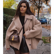 Donna con cappotto beige oversize, borsa a tracolla marrone, maglia nera e gonna in pelle. Moda autunnale, look urbano elegante.