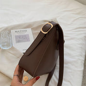 Borsa a tracolla marrone in pelle su letto bianco, con fibbia dorata. Accanto, libro "Matisse" e bicchiere d'acqua. Elegante accessorio moda.