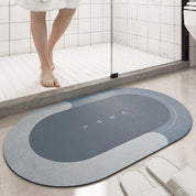 Tappeto da bagno ovale blu con scritta "HOME", antiscivolo, posizionato davanti a una doccia moderna, accanto a pantofole bianche.