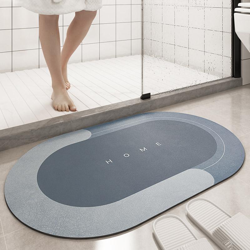 Tappeto da bagno ovale blu con scritta "HOME", antiscivolo, posizionato davanti a una doccia moderna, accanto a pantofole bianche.