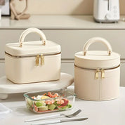 Due eleganti borse termiche beige con zip dorata su tavolo da cucina, accanto a contenitore di vetro con insalata fresca. Accessori pranzo moderni.