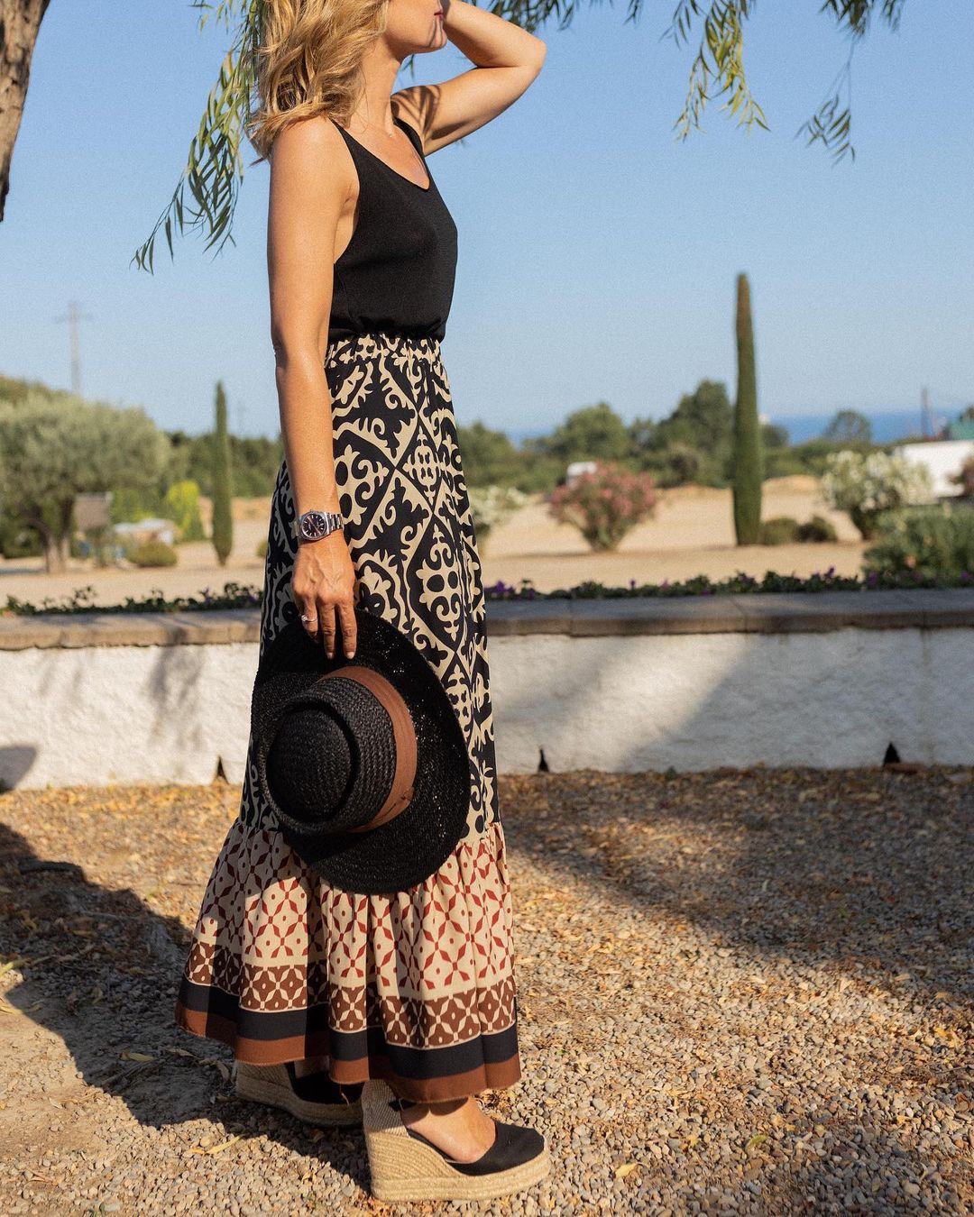 Donna con abito lungo boho nero e beige, cappello di paglia in mano, sandali con zeppa, in giardino soleggiato. Moda estiva elegante.