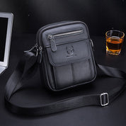 Borsa a tracolla in pelle nera su sfondo scuro, con cerniere e logo, accanto a un bicchiere di whisky e un laptop. Elegante e funzionale.