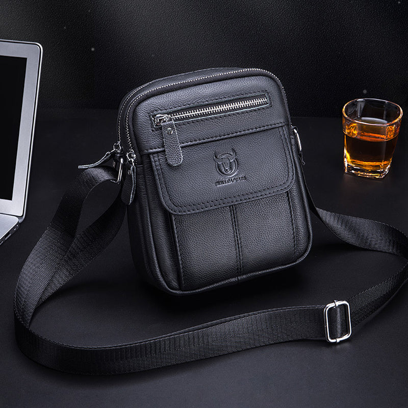 Borsa a tracolla in pelle nera su sfondo scuro, con cerniere e logo, accanto a un bicchiere di whisky e un laptop. Elegante e funzionale.