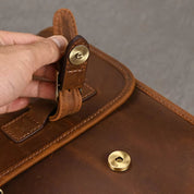 Mano che apre una borsa in pelle marrone con chiusura a bottone dorato. Accessorio elegante, ideale per lavoro e viaggi.