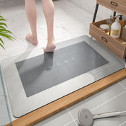 Tappetino da bagno antiscivolo grigio con scritta "HOME", design moderno, su pavimento in marmo, accanto a mobile in legno con accessori da bagno.