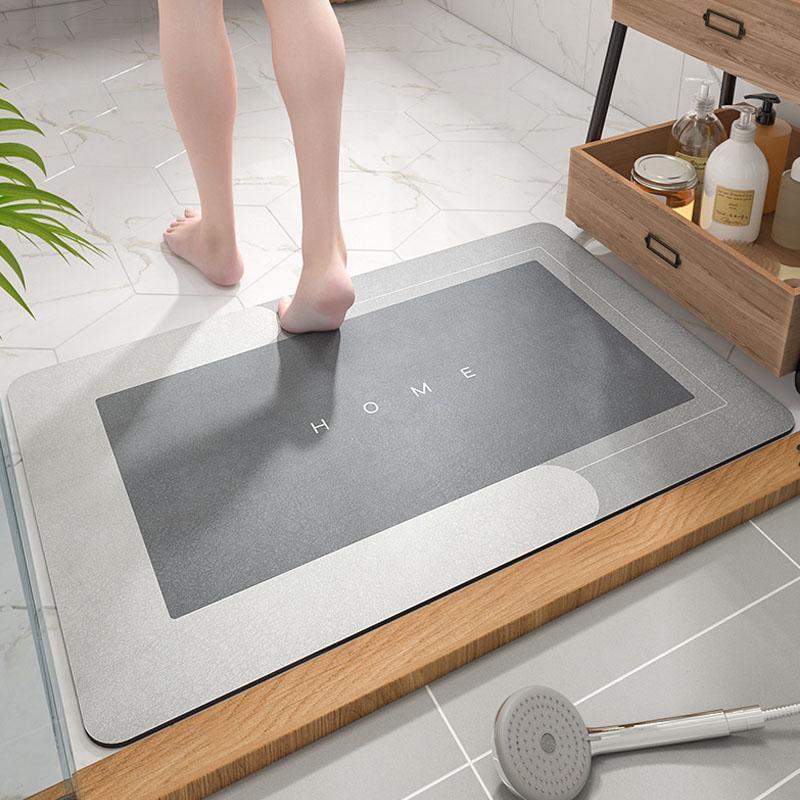 Tappetino da bagno antiscivolo grigio con scritta "HOME", design moderno, su pavimento in marmo, accanto a mobile in legno con accessori da bagno.