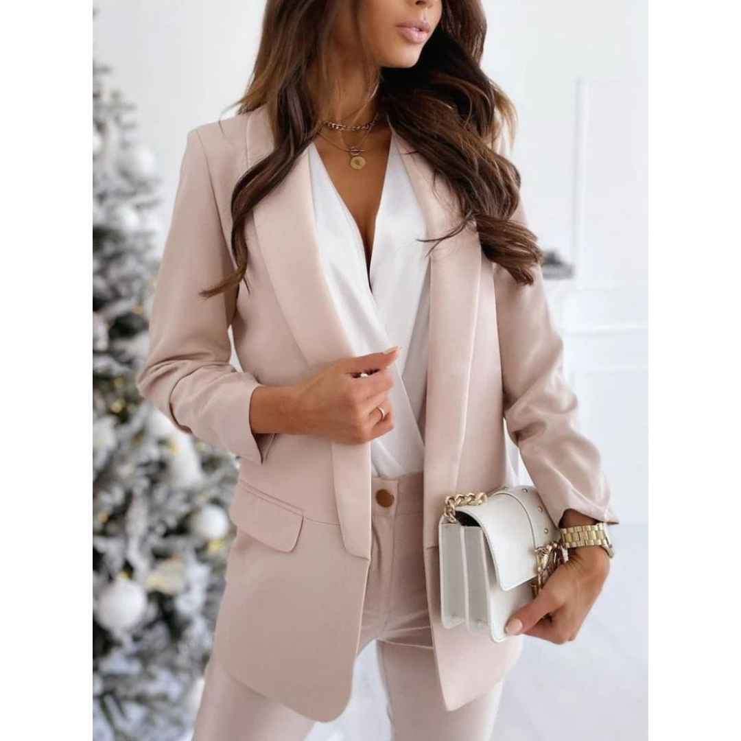 Donna in elegante completo blazer rosa chiaro, camicia bianca, borsa a tracolla bianca, davanti ad albero di Natale decorato. Moda femminile chic.