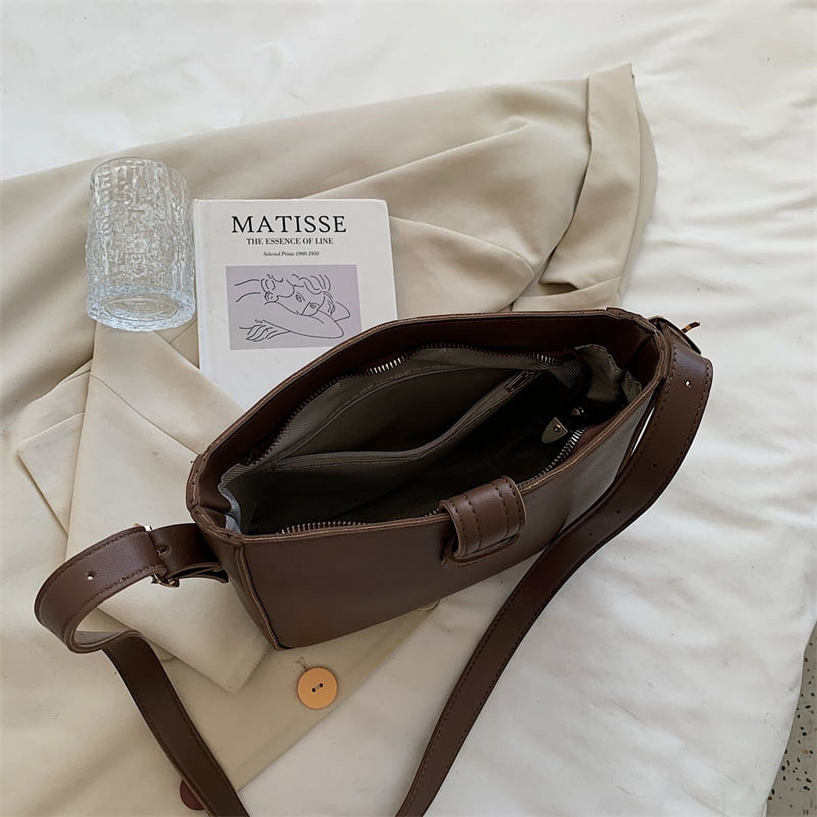 Borsa a tracolla marrone in pelle su letto bianco, accanto a libro "Matisse", bicchiere d'acqua e camicia beige. Accessori moda eleganti.