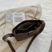 Borsa a tracolla marrone in pelle su letto bianco, accanto a libro "Matisse", bicchiere d'acqua e camicia beige. Accessori moda eleganti.