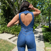 Donna con tuta in denim blu con schiena a cuore, passeggia su marciapiede alberato. Moda estiva, abbigliamento casual, stile urbano.