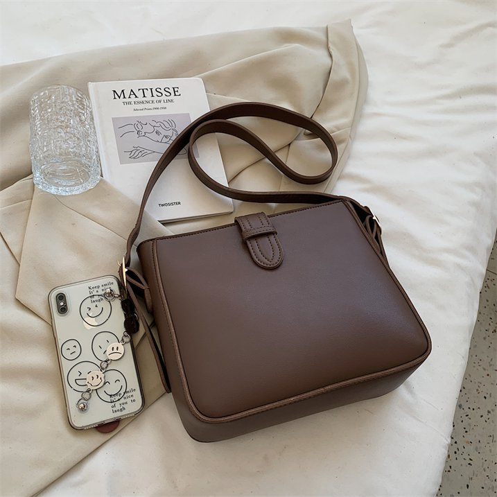 Borsa a tracolla marrone su letto bianco, accanto a libro "Matisse", bicchiere d'acqua e smartphone. Accessorio moda elegante e versatile.