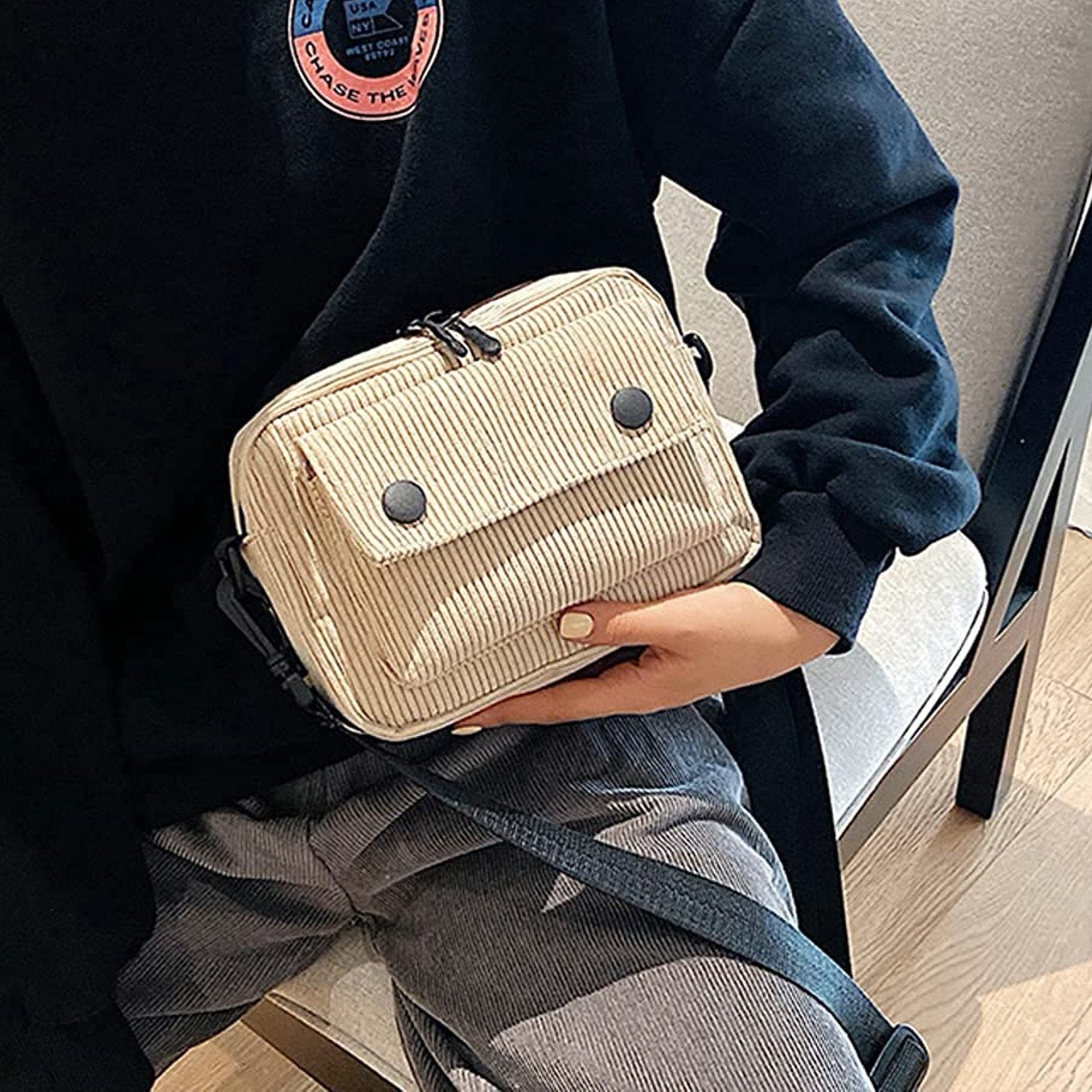 Borsa a tracolla beige in tessuto a coste, design compatto con doppia zip e bottoni decorativi, indossata da una persona seduta su una sedia.