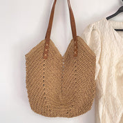 Borsa a tracolla in paglia intrecciata con manici in pelle marrone, design estivo, ideale per spiaggia o shopping, appesa accanto a camicia bianca.