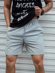 Uomo in pantaloncini sportivi grigi e maglietta nera davanti a una porta bianca. Abbigliamento casual estivo, moda maschile.