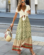 Donna sorridente con occhiali da sole bianchi, indossa una gonna lunga verde con motivi geometrici e una camicetta bianca annodata, borsa di paglia.