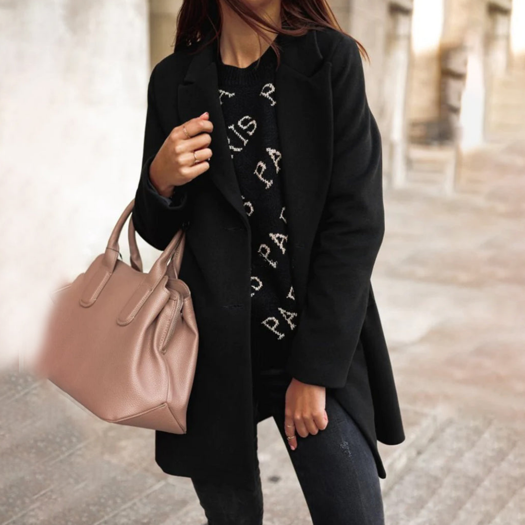 Donna con cappotto nero elegante, maglione con scritta "Paris", jeans scuri e borsa a mano beige. Moda autunnale, stile urbano chic.