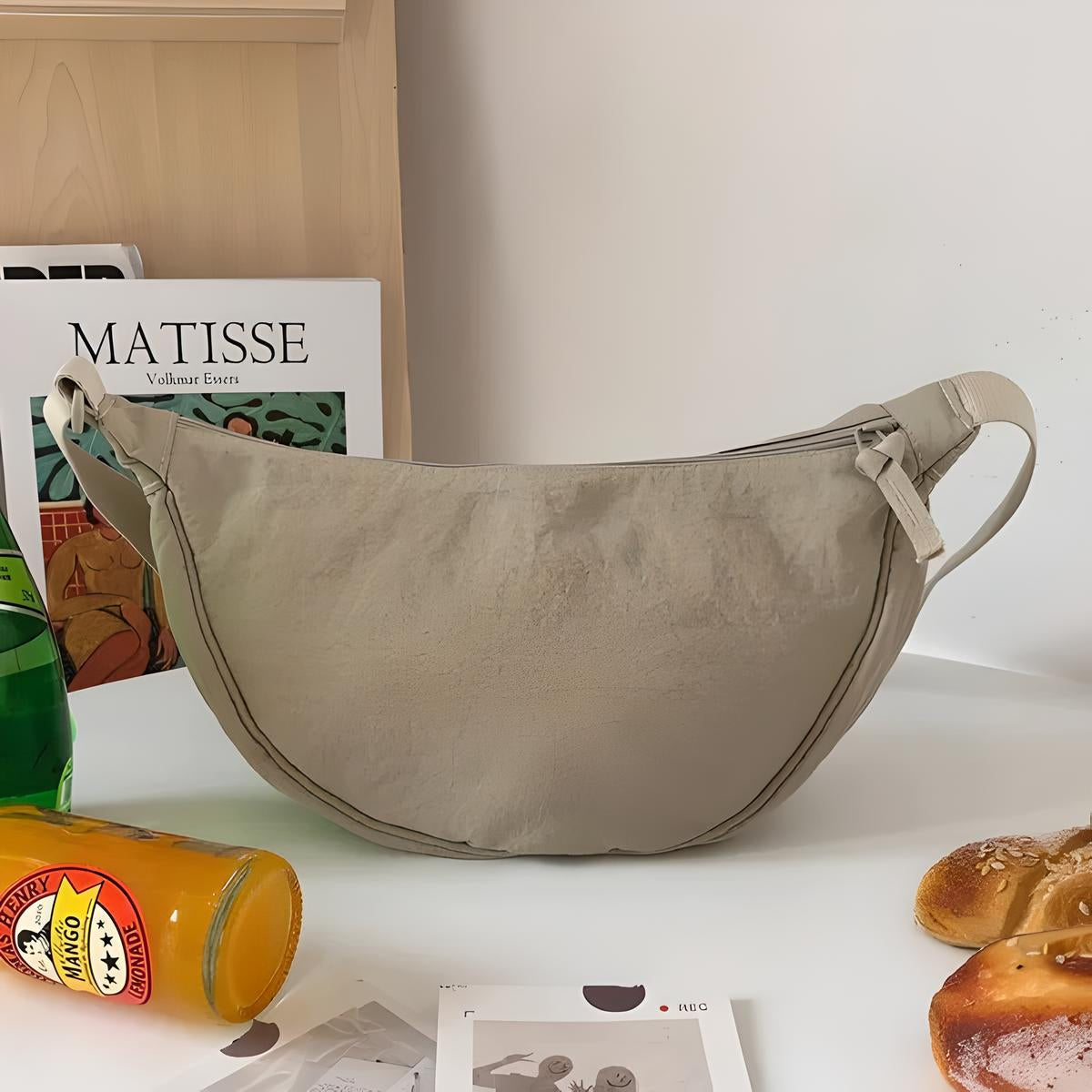 Borsa a tracolla beige su tavolo bianco, accanto a riviste, bottiglia di succo e pane. Design minimalista, ideale per moda casual e quotidiana.