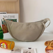 Borsa a tracolla beige su tavolo bianco, accanto a riviste, bottiglia di succo e pane. Design minimalista, ideale per moda casual e quotidiana.