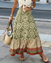 Gonna lunga boho verde con motivi geometrici, abbinata a borsa di paglia e sandali. Moda estiva donna, stile casual chic.