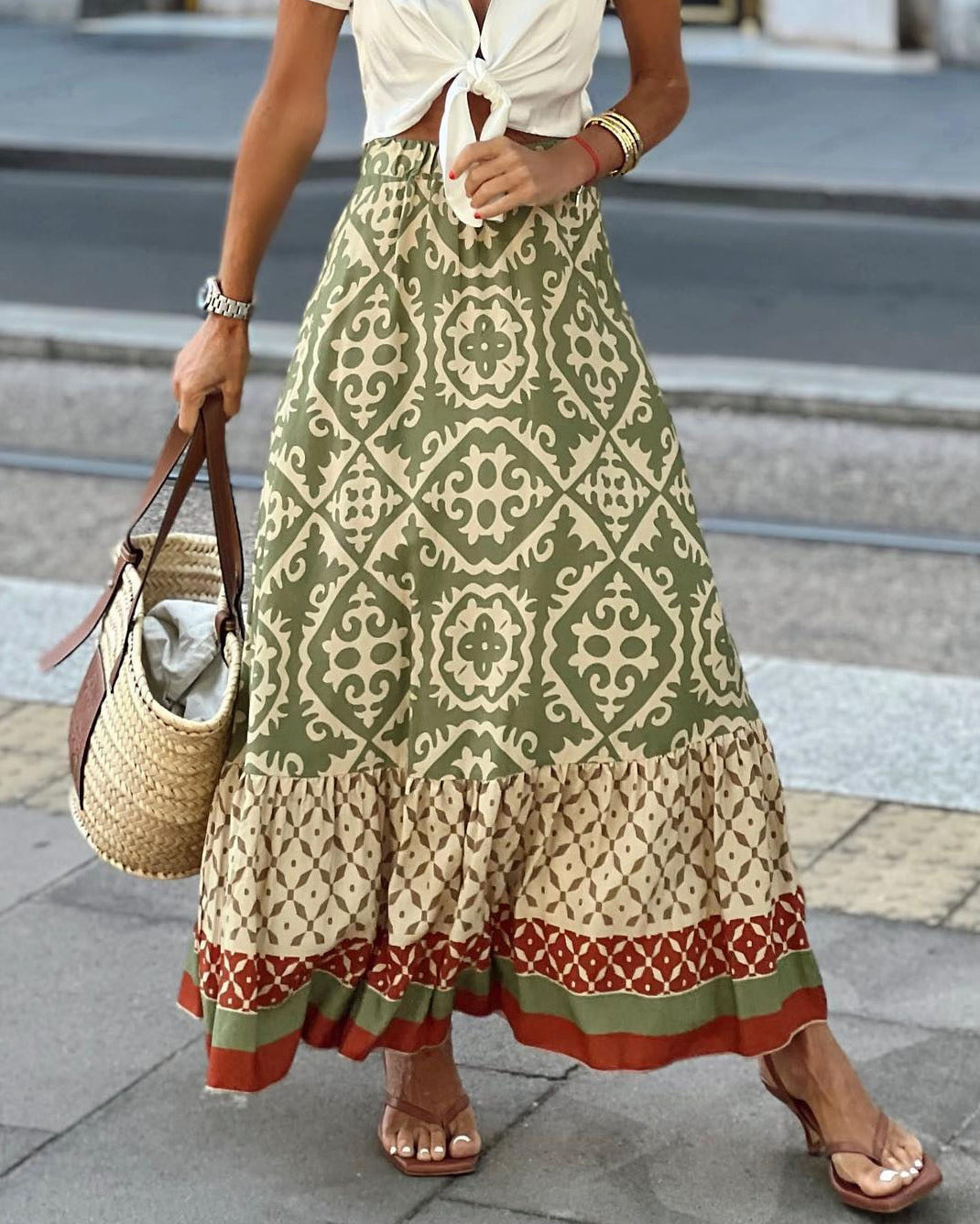 Donna con gonna lunga boho verde e beige, motivo geometrico, top bianco annodato, sandali marroni, borsa di paglia. Moda estiva casual chic.