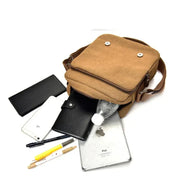 Borsa a tracolla marrone in tela con tasche multiple, contenente bottiglia d'acqua, iPad, smartphone, portafoglio nero e penne. Accessori moda uomo.