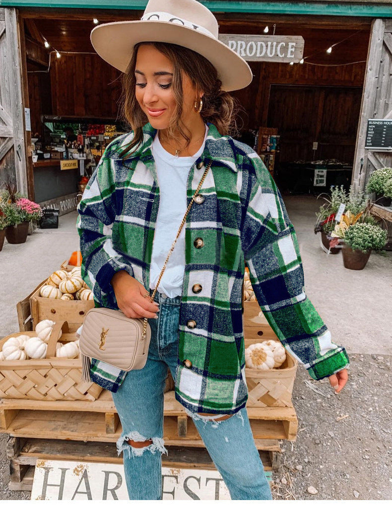 Donna con cappello beige e camicia a quadri verde e blu, davanti a un mercato agricolo. Indossa jeans strappati e borsa a tracolla.