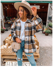 Donna con cappello beige e giacca a quadri marrone, bianca e nera, davanti a un mercato agricolo. Indossa jeans strappati e borsa a tracolla.