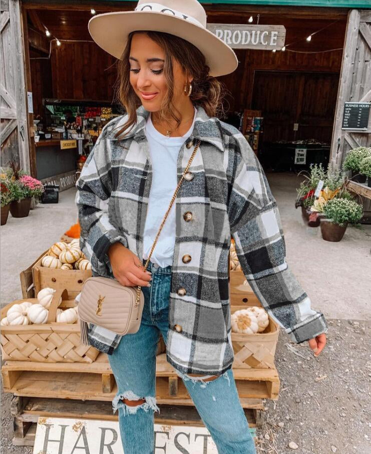 Donna con cappotto a quadri grigio, jeans strappati e borsa beige, davanti a un mercato agricolo con zucche e fiori. Moda autunnale casual.