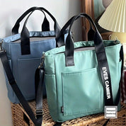 Due borse a tracolla impermeabili in tessuto blu e verde con manici neri, tasche frontali e dettagli moderni, su un tavolo di vimini.