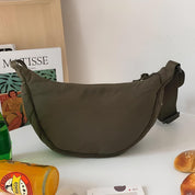 Borsa a tracolla verde oliva su tavolo bianco, accanto a libro d'arte, bottiglia di vetro e panini. Design moderno, ideale per moda casual.