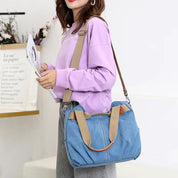 Donna con borsa a tracolla in tela blu, manici beige, indossa maglia viola e pantaloni grigi. Accessorio moda casual, stile urbano.