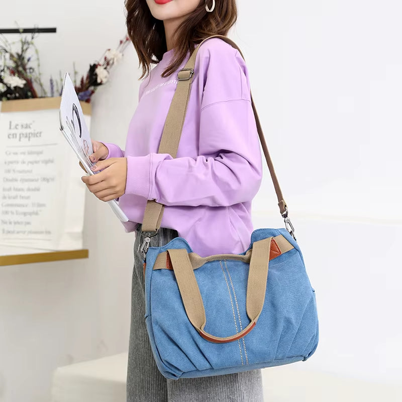 Donna con borsa a tracolla in tela blu, manici beige, indossa maglia viola e pantaloni grigi. Accessorio moda casual, stile urbano.