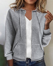 Giacca donna grigia con cappuccio, tessuto a trama waffle, zip frontale, tasche laterali, indossata su maglietta bianca. Moda casual autunnale.