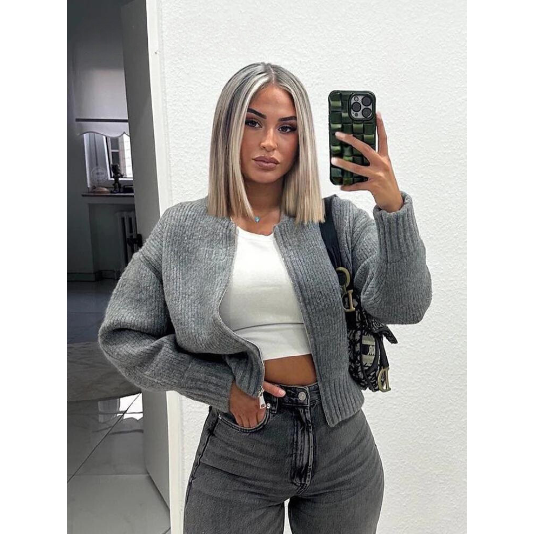 Donna con capelli biondi e lisci, indossa cardigan grigio, top bianco e jeans. Scatta selfie con smartphone in una stanza moderna.