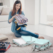 Donna sorridente piega vestiti colorati accanto a organizer da viaggio in tessuto azzurro, seduta su pavimento di soggiorno moderno e luminoso.