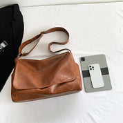 Borsa a tracolla in pelle marrone su letto bianco, accanto a tablet e smartphone. Accessori moda eleganti, design moderno, stile casual.