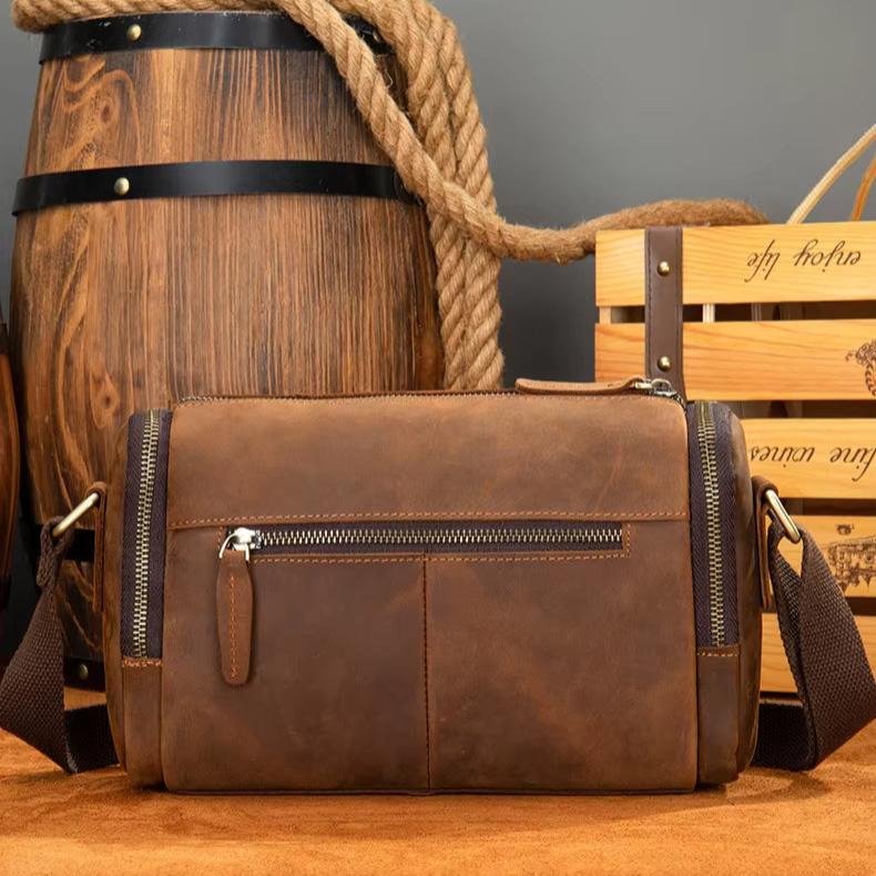 Borsa a tracolla in pelle marrone vintage, con cerniere e tasche frontali, su sfondo rustico. Accessorio moda uomo elegante e pratico.